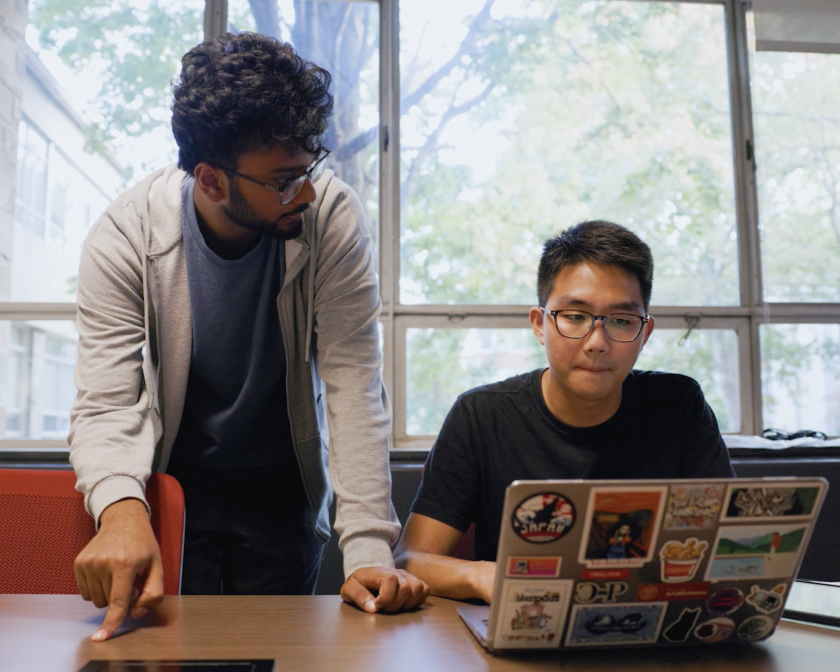 two students (one standing, one sitting) look at computer laptop